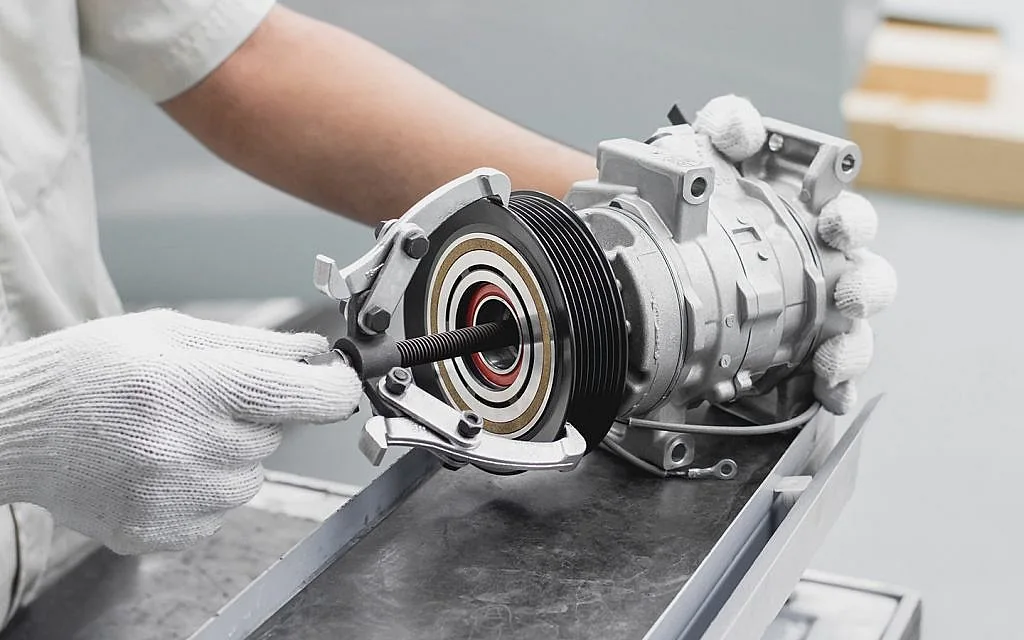 Gloved hands holding a pulley tool against a removed AC compressor